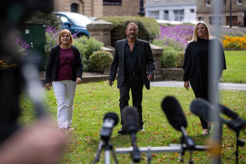 Three people walk towards a press conference and awaiting cameras.