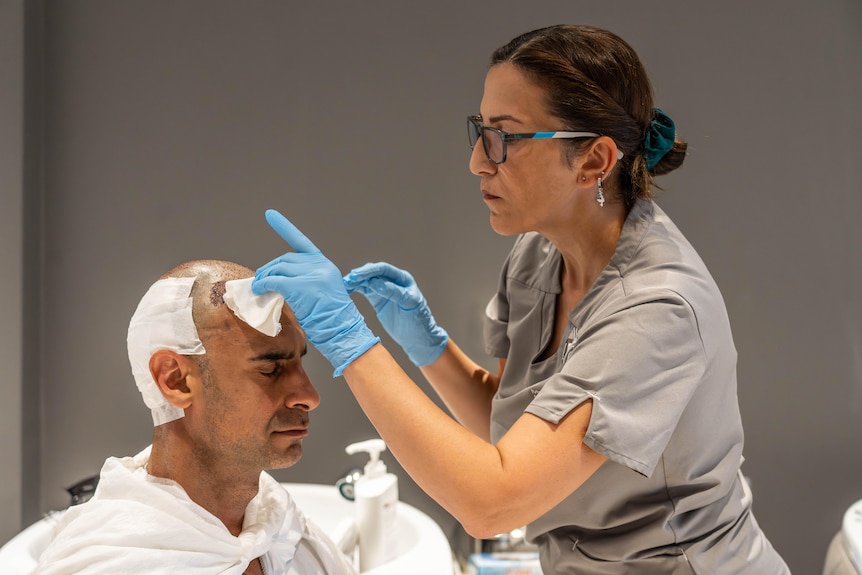 A nurse wearing blue surgical gloves dabs a wound on a man's scalp, grimaces slightly with closed eyes 