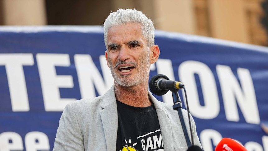 A man with white hair speaks at a refugee rally with a blue banner behind him.