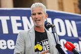 A man with white hair speaks at a refugee rally with a blue banner behind him.