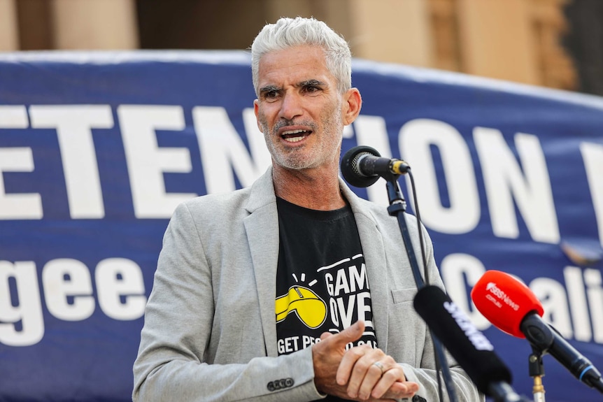 A man with white hair speaks at a refugee rally with a blue banner behind him.