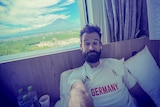 A man in a Germany jersey lying in a hotel room bed with a mountain view from his window 