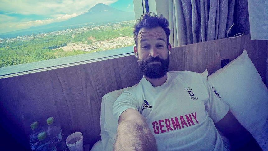 A man in a Germany jersey lying in a hotel room bed with a mountain view from his window 