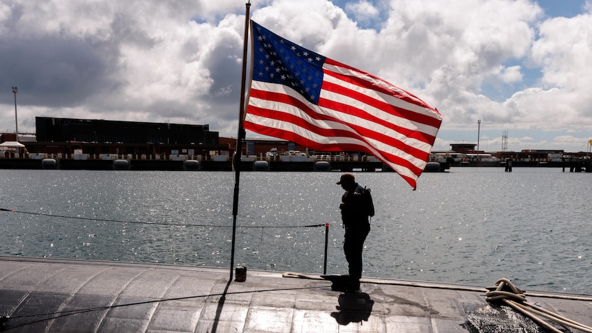 A ship crew member stands on a submarine with the US flag in the background