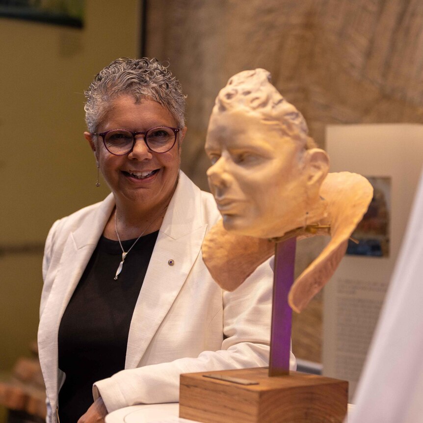 Deborah Cheetham Fraillon stands next to a bust of her face with a high collar in ochre-colored clay. She smiles at the camera.