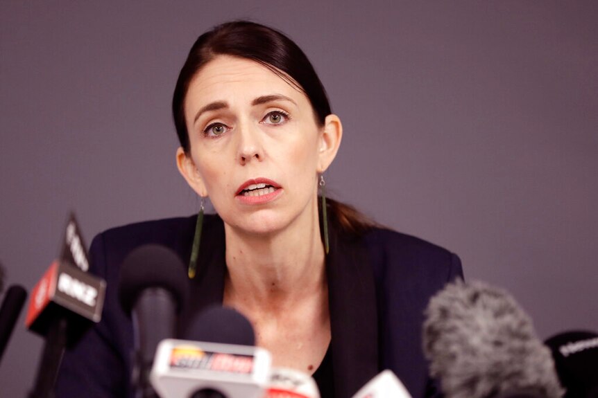Jacinda Ardern sits in front of a warm grey backdrop in front of a string of media microphones.