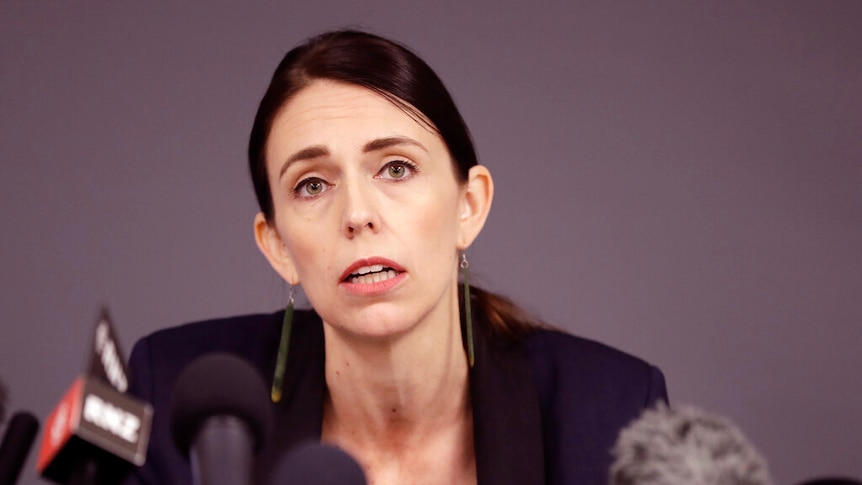 Jacinda Ardern sits in front of a warm grey backdrop in front of a string of media microphones.