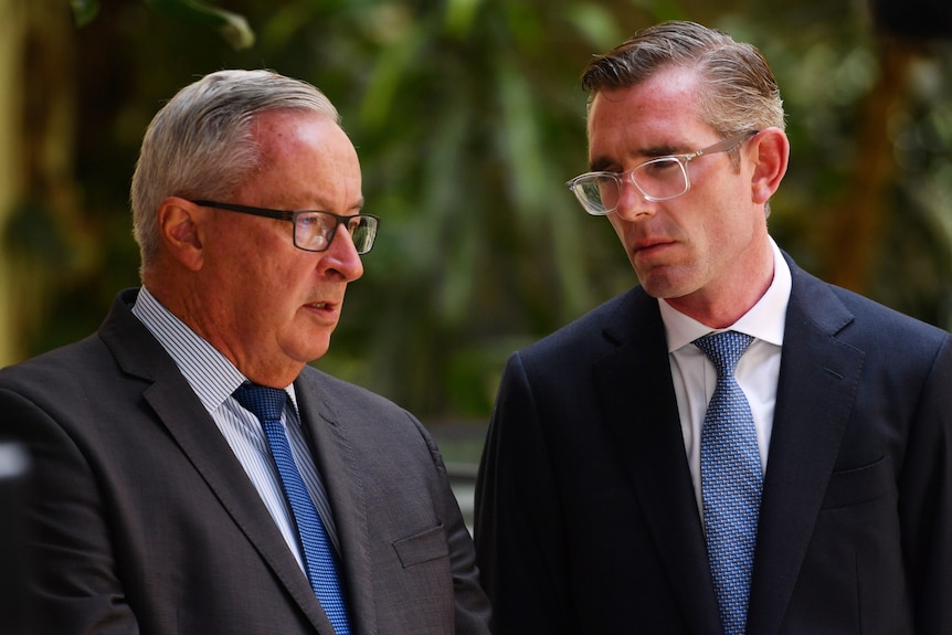 Brad Hazzard  and Dominic Perrottet, both wearing dark suits, blue ties, stand next to each other talking, greenery behind.