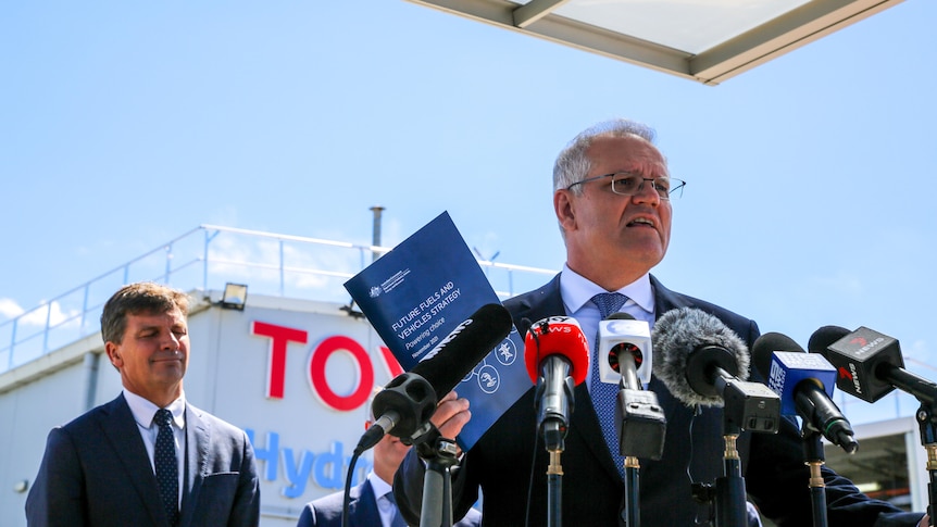 Morrison holds up a printed policy paper in front of a bank of microphones