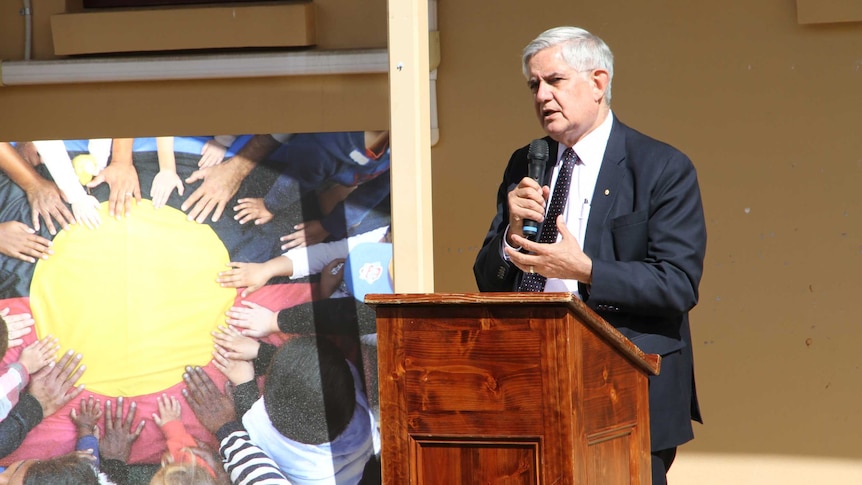 Ken Wyatt stands with a microphone next to a canvas of a photo with different coloured hands touching an Aboriginal flag