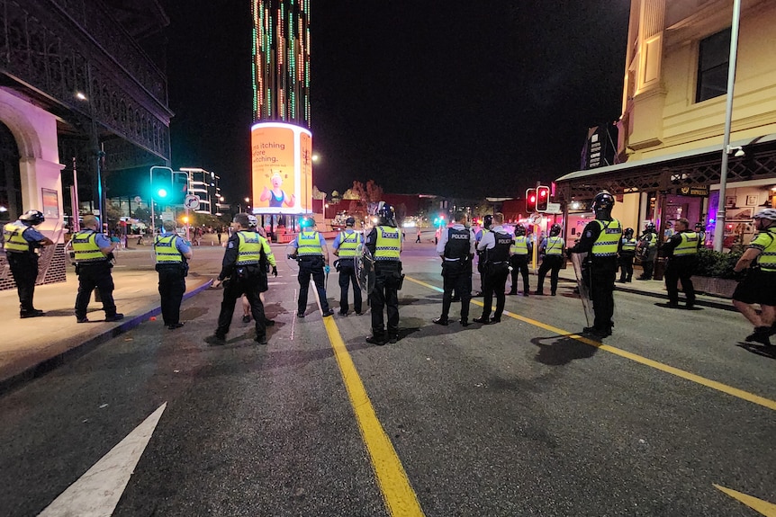 A line of police in riot gear stands across a road in Perth's CBD near the train station at night.