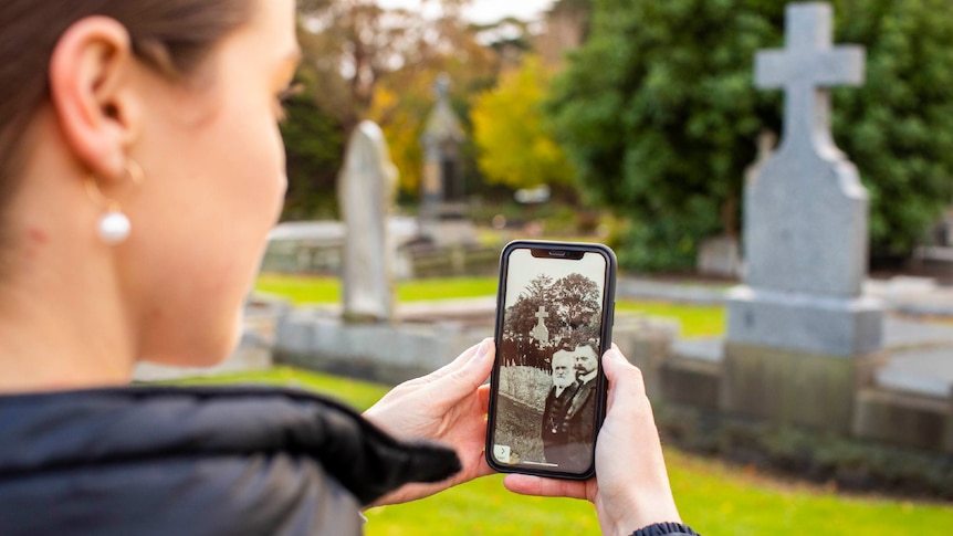 A woman uses an augmented reality app and points her phone at a grave. Two men in Victorian clothing appear on screen.
