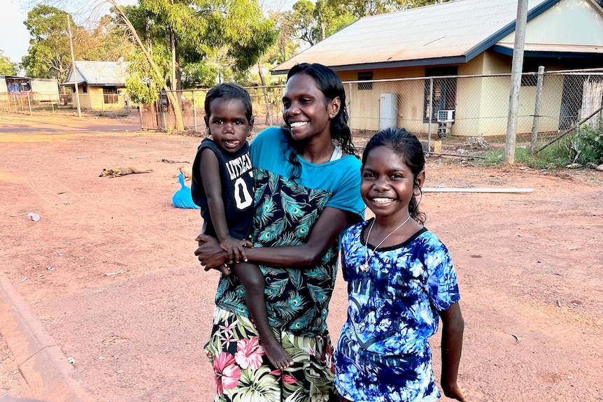 A mum with two young kids outside a house, smiling.