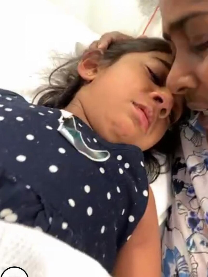 Tharnicaa laying on a hospital bed with her mother holding her head and comforting her.