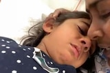 Tharnicaa laying on a hospital bed with her mother holding her head and comforting her.