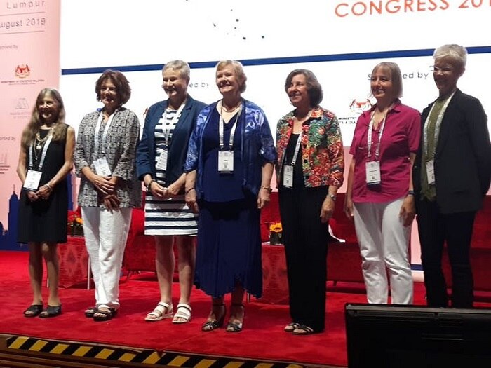Seven women stand in a row for the camera with a conference banner behind them.