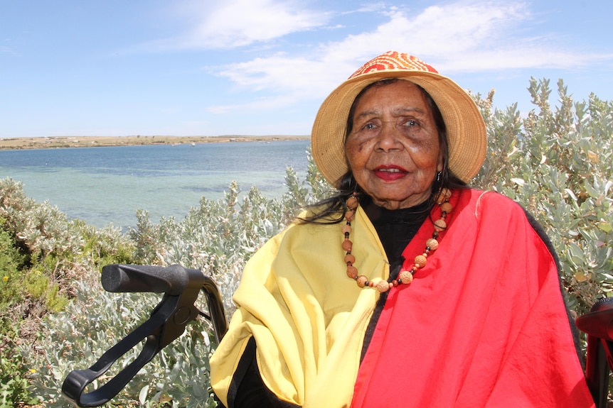 An elderly Indigenous woman wears a hat, necklace around neck, cream, red and black top, sits on walker, sea, shrub behind her.