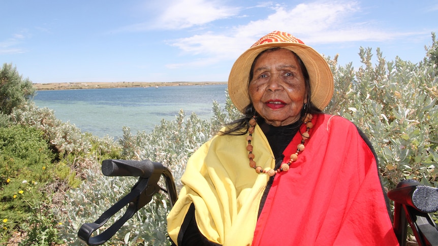 An elderly Indigenous woman wears a hat, necklace around neck, cream, red and black top, sits on walker, sea, shrub behind her.