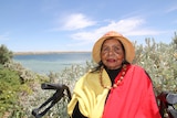 An elderly Indigenous woman wears a hat, necklace around neck, cream, red and black top, sits on walker, sea, shrub behind her.