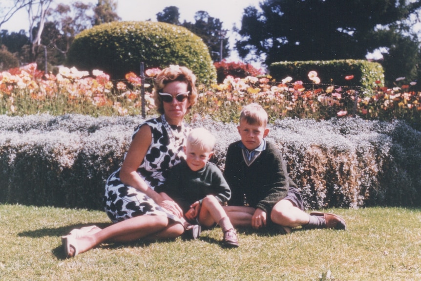 A woman wearing a dress and sunglasses sits in a garden with two young boys with blonde hair