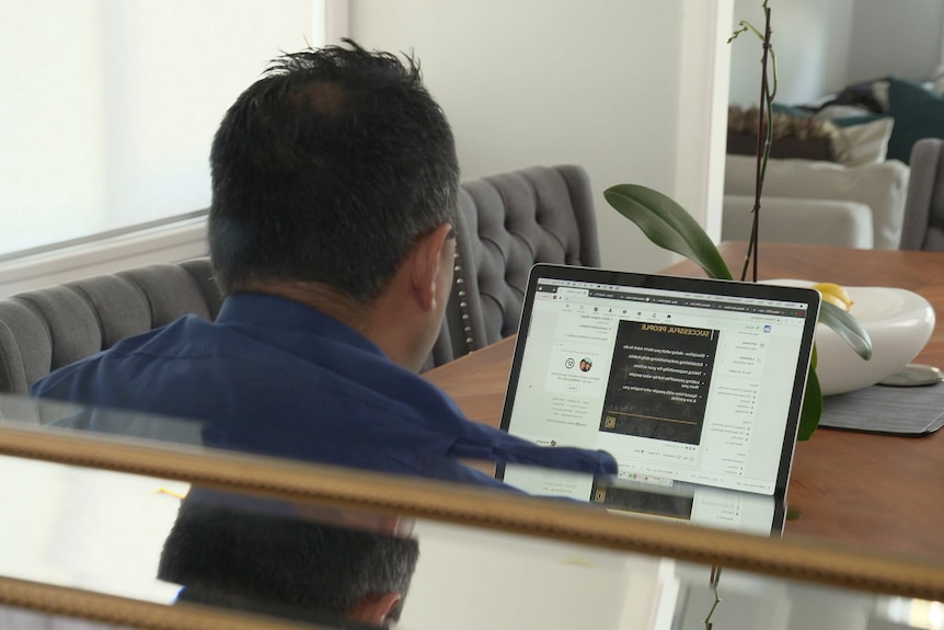 A man sitting behind a computer at a dining room table at home.
