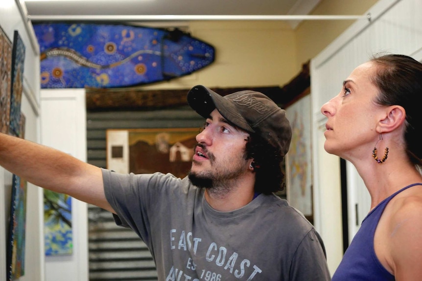 Aboriginal artist points to art and describes dream time story to patron.