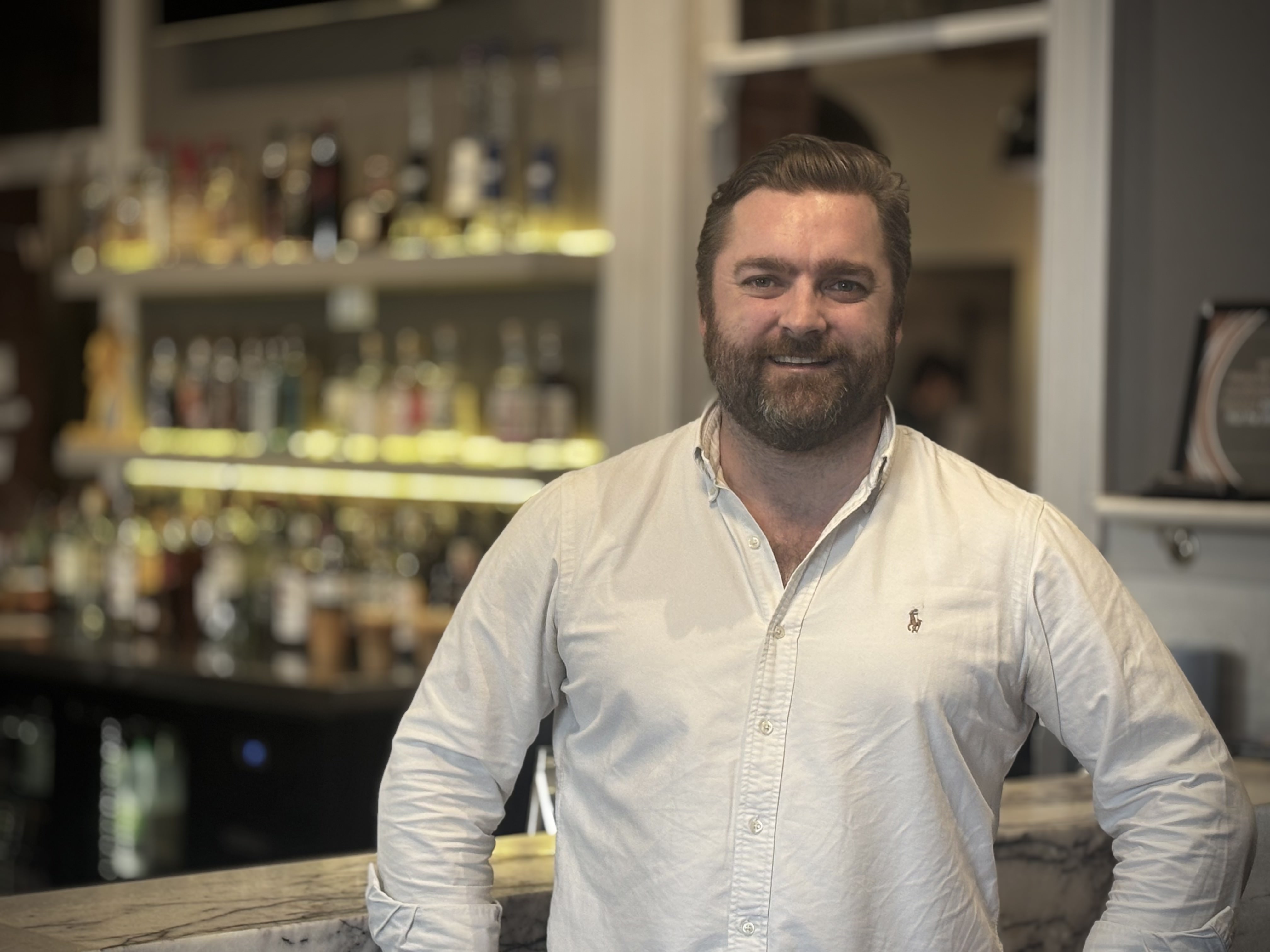 Um homem com barba e cabelo castanhos vestindo uma camisa branca em frente a um bar