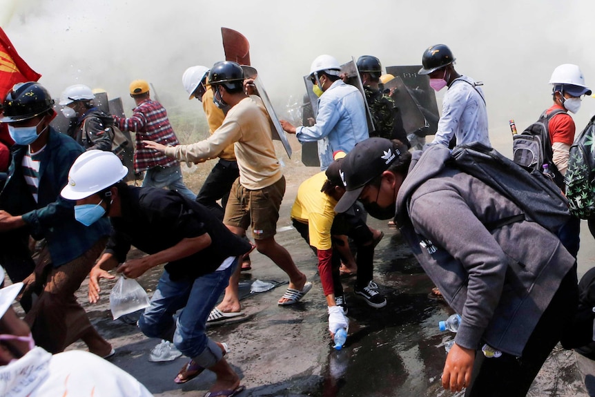 A chaotic street scene shows protesters wearing helmets and face masks running from tear gas attack.