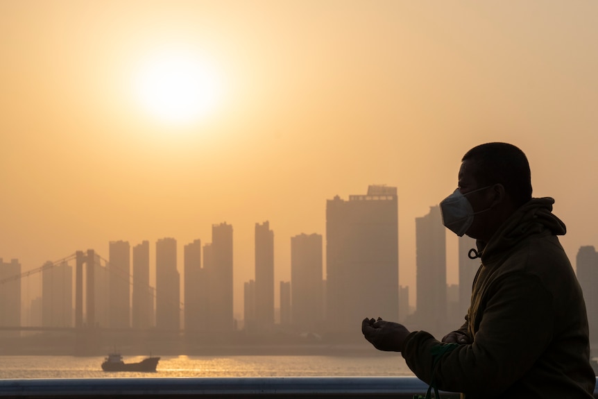 A man stands on a river bank with skyscrapers in the background.