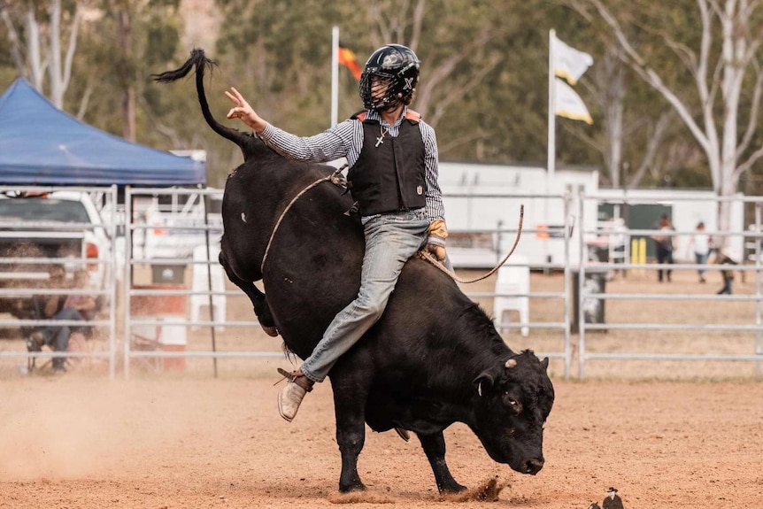 A teenager sits on top of a bucking bull in an open-air rodeo arena