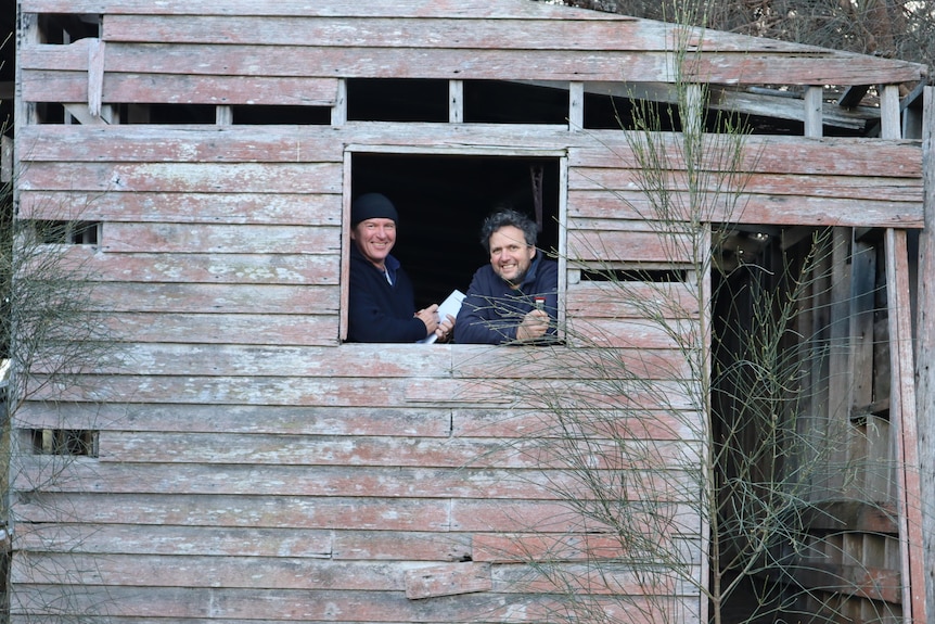 Two men stand in a window smiling. They are in a building in need of repair