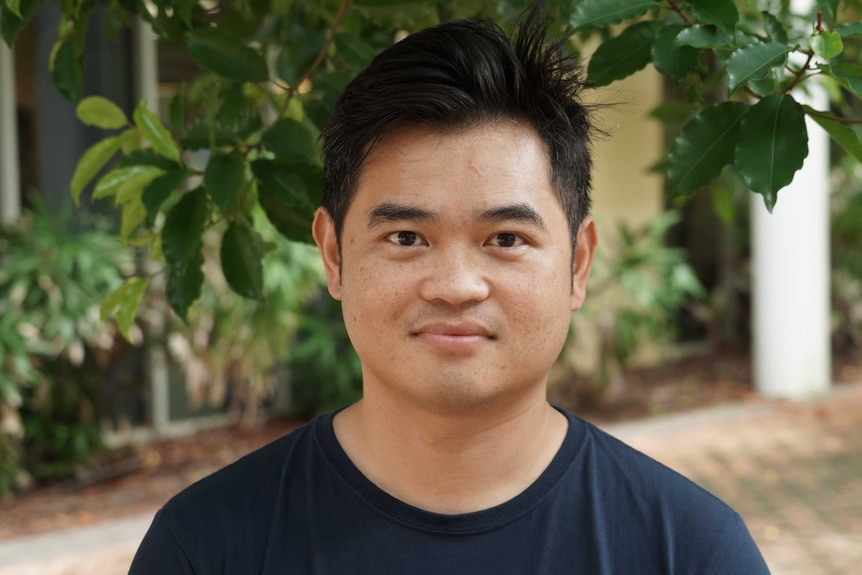 A young man stands in front of a camera and smiles faintly.