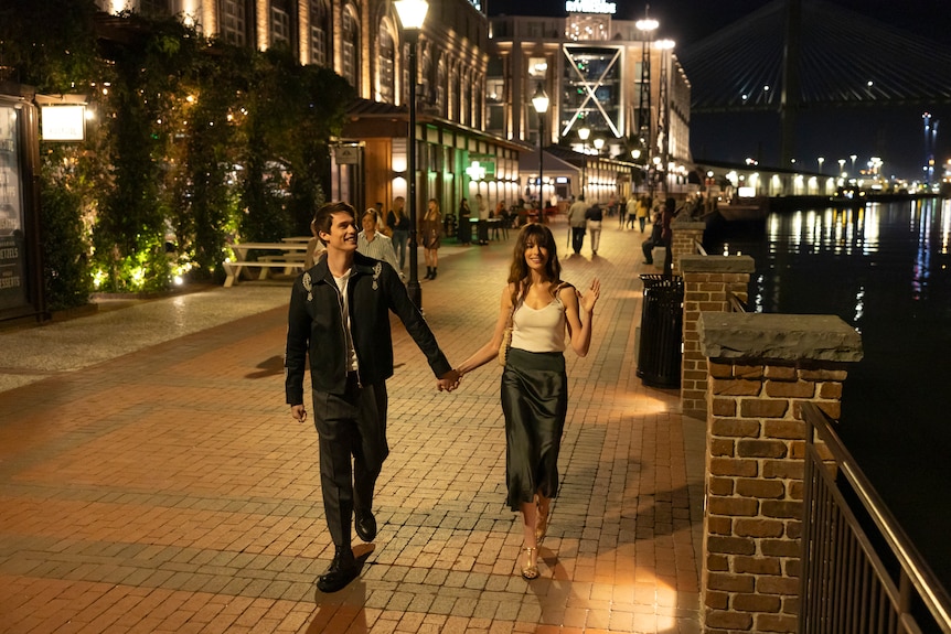 A film still of Nicholas Galitzine, 29, and Anne Hathaway, 41, smiling as they hold hands while walking by a riverside at night.