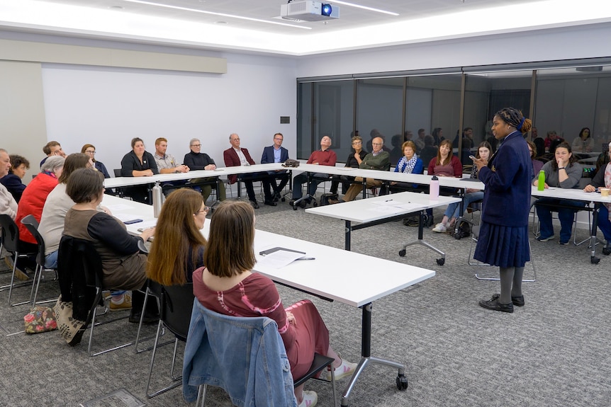 Une femme debout parle à une salle de personnes assises à des tables.