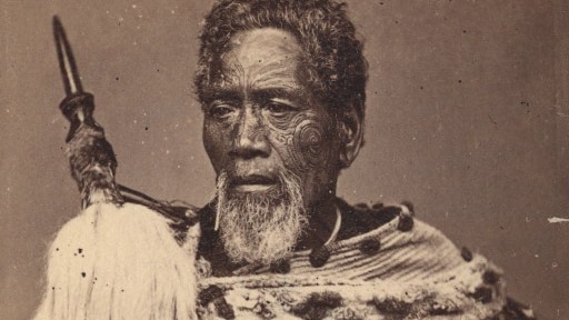 A sepia photo circa 1800s of a Maori chief in traditional dress holding a traditional spear-like object.