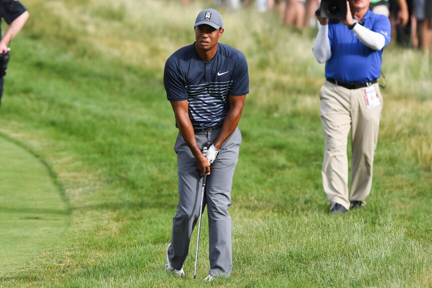 Tiger Woods looks into the distance standing in the rough with his golf club between his legs.