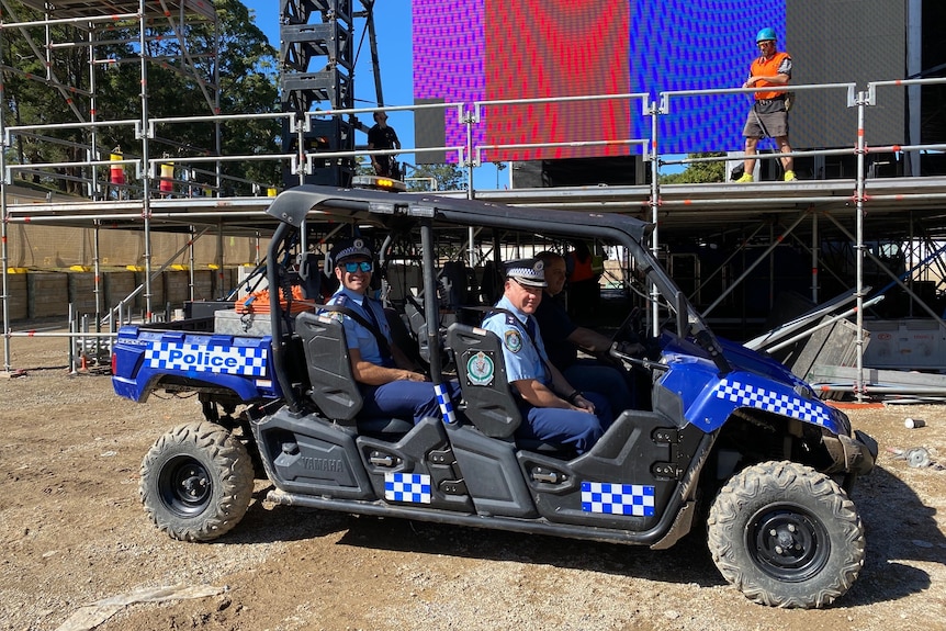 Two police officers sit in a blue police buggy in front of a music festival stage