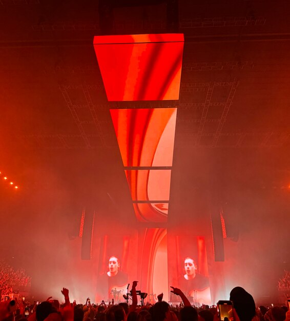 Red lighting shows a concert with large screens in front of a crowd and ceiling screens with red and orange swirls