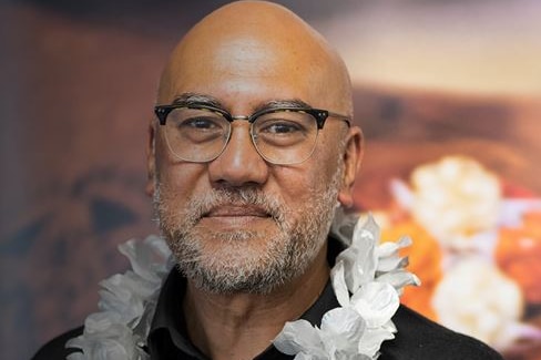 Man wears frames and black top, stares to camera and wears white flower lei around his neck.