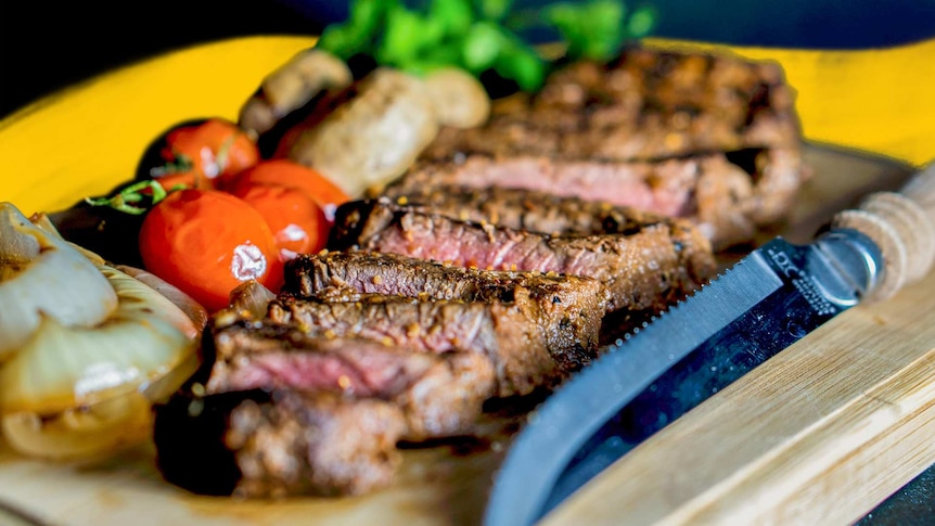 An image of cooked steak, sliced, on a board with bright vegetables.
