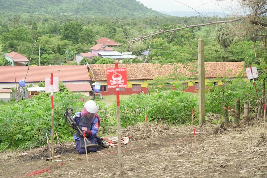 一名男子跪在地上，手持探测器，位于俯瞰建筑物的山丘上。