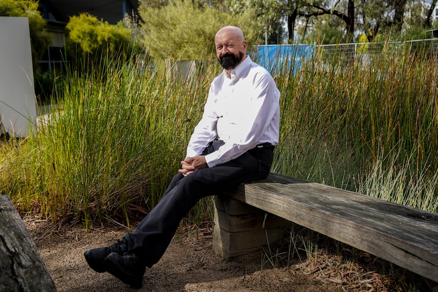 A man with a black beard sits for a portrait in a university garden