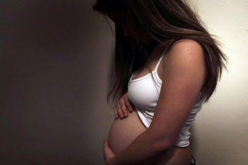 A pregnant woman in a white top holds her belly.