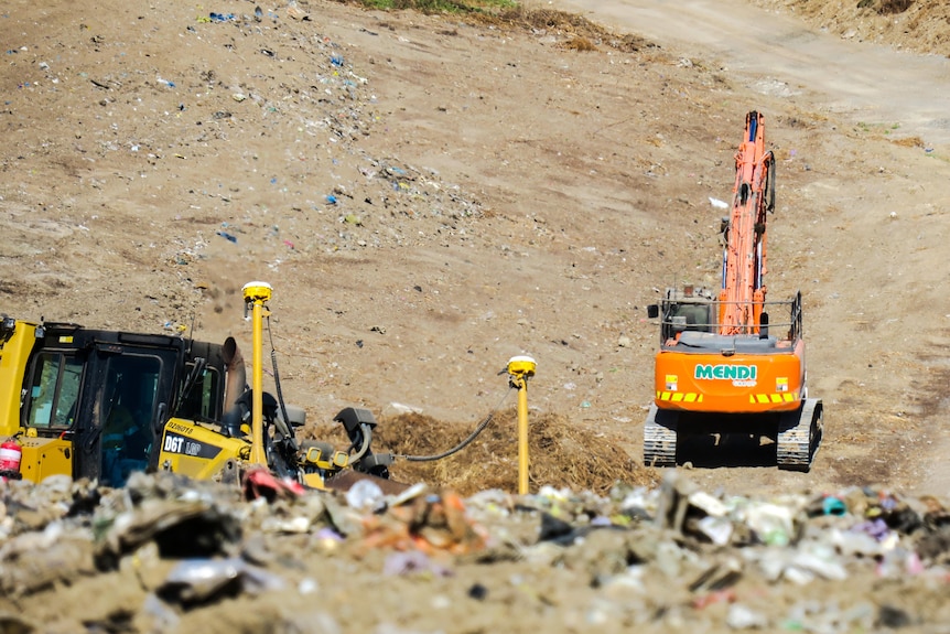 A mound of waste sits in the foreground with plastic bags sticking through the dirt, bulldozers clear land in the background.