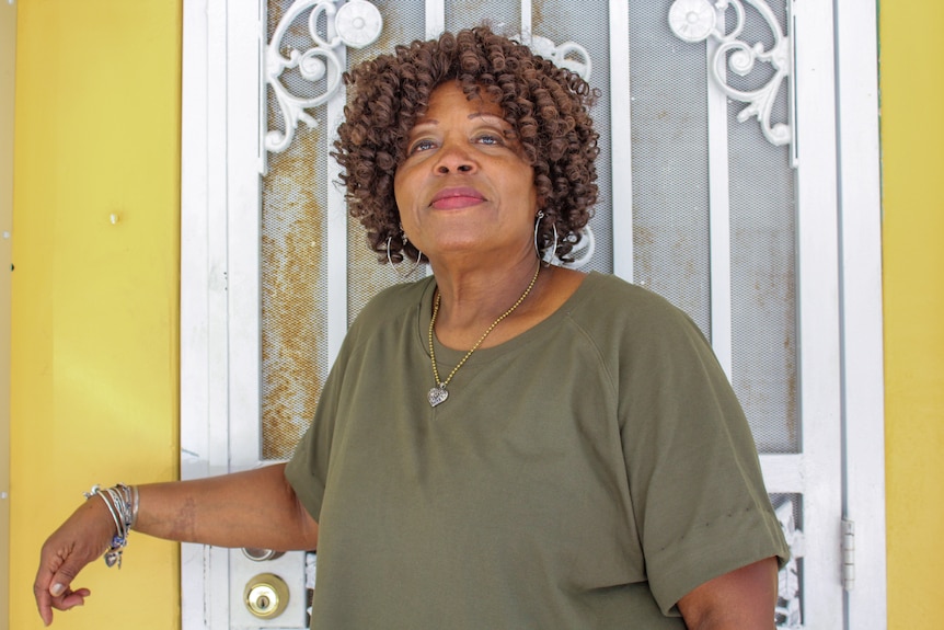 A Black woman in a green shirt poses for a portrait in front of a yellow house