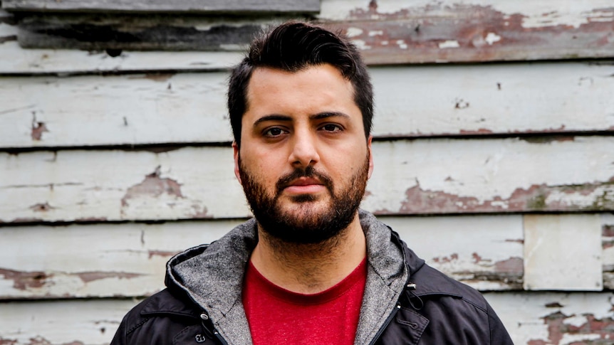 Poet Omar Sakr stands in front of a chipped and crumbling weatherboard wall.