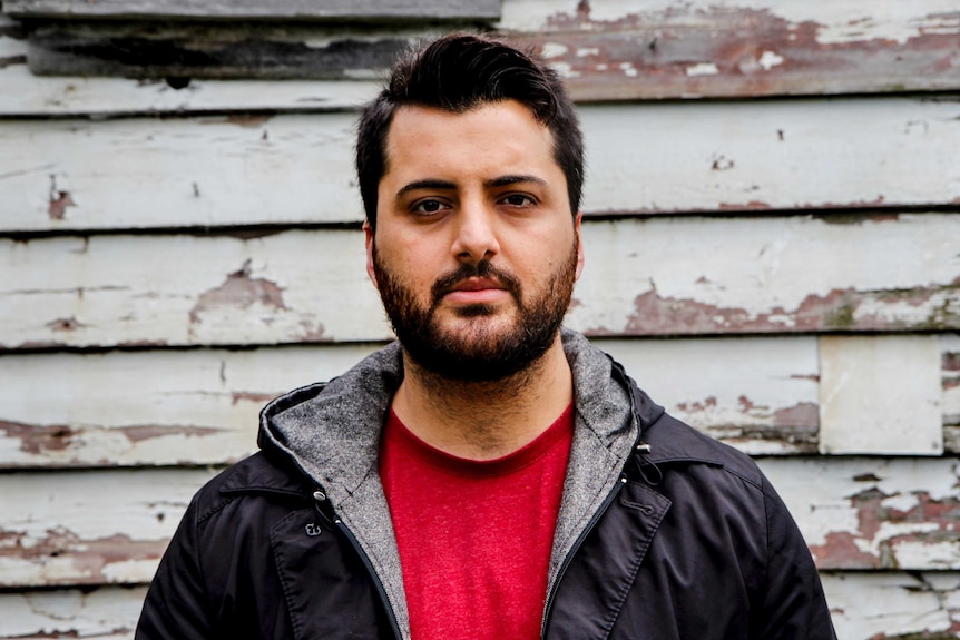 Poet Omar Sakr stands in front of a chipped and crumbling weatherboard wall.