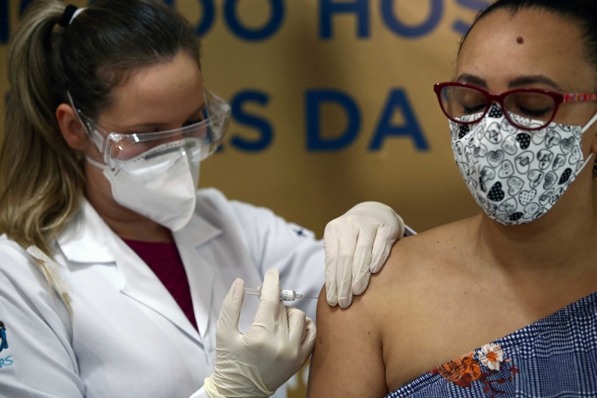 O tânără femeie care poartă o mască de față închide ochii în timp ce o asistentă mascată o injectează cu o seringă.