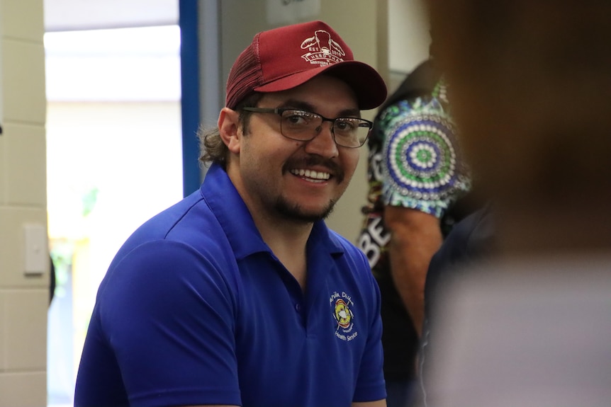 A young man with a moustache and glasses wearing a red cap smiles at the camera