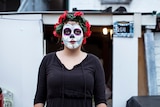 A woman with a painted face for Day of the Dead stands in front of a house.